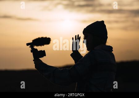 Vue latérale d'une jeune femme blogueuse méconnue dans un chapeau souriant et montrant la main tout en filant sur un appareil photo numérique contre un soleil Banque D'Images