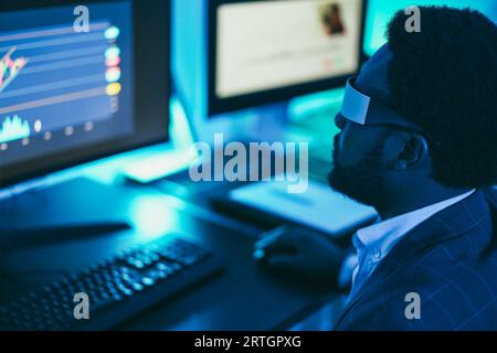 Trader africain travaillant à l'intérieur du bureau la nuit portant un casque de réalité virtuelle - concept de trading et de technologie - Soft focus sur les lunettes Banque D'Images