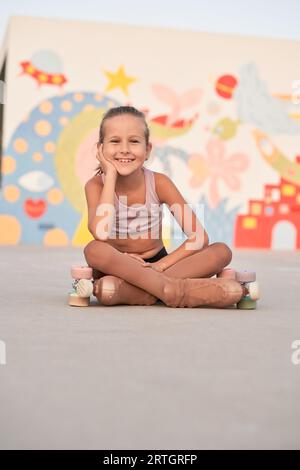 Portrait de souriante préadolescente en vêtements d'activité et patins à roulettes quad assis sur un terrain de sport avec les jambes croisées et regardant la caméra Banque D'Images