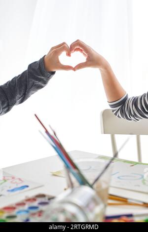 Mains des enfants anonymes de récolte montrant le geste en forme de cœur à la table blanche avec des peintures colorées et des pinceaux dans le studio de lumière Banque D'Images