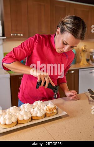 Chef pâtissier féminin en uniforme rose brûlant meringue suisse avec torche à gaz pour la pâtisserie Banque D'Images