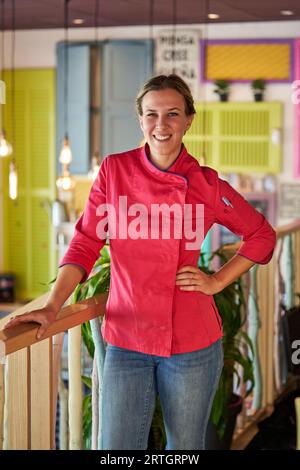 Souriant chef féminin confiant dans des vêtements professionnels avec la main sur la taille debout près de la balustrade en bois dans la pièce colorée et regardant la caméra Banque D'Images