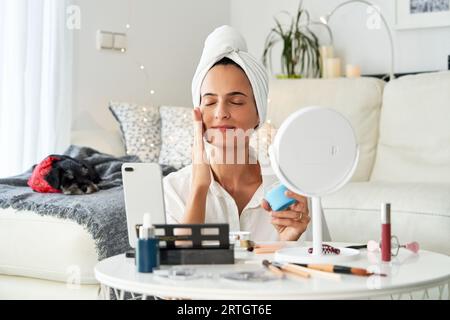 Femme ravie blogueuse beauté appliquant la crème hydratante rajeunie sur le visage tout en streaming vidéo pour vlog Banque D'Images