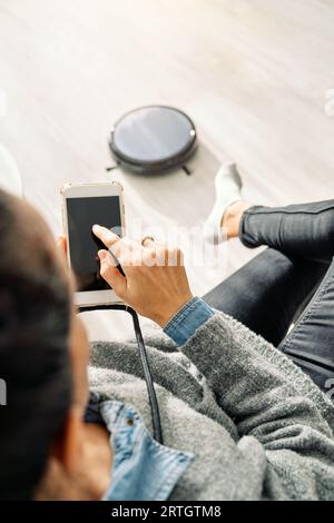 D'en haut de la récolte méconnaissable femme assise sur le canapé et la navigation smartphone tandis que l'aspirateur robotique moderne travaille sur le sol dans le salon A. Banque D'Images