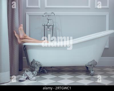 Femme pieds nus méconnaissable couchée dans une baignoire blanche avec les jambes dehors et croisée dans la salle de bains de l'hôtel Banque D'Images