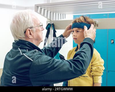 Vue latérale de l'instructeur masculin âgé mettant sur le bandeau de sport sur la tête de l'enfant tout en se préparant pour la leçon de tennis dans le vestiaire Banque D'Images