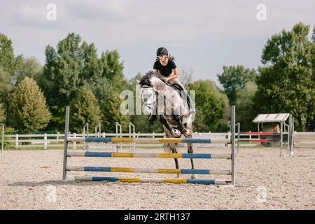 Une jeune fille à bord d'un cheval gris pommier sautant sur un obstacle au manège du centre équestre par une journée ensoleillée Banque D'Images