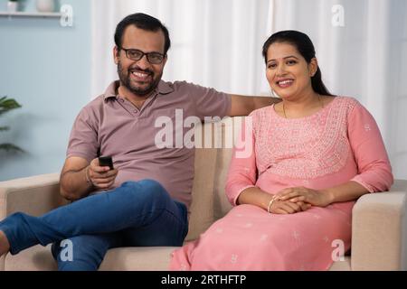 Heureux couple indien d'âge moyen détendu regardant la télévision ou la télévision en se parlant sur le canapé à la maison - concept de divertissement, de lien familial et Banque D'Images