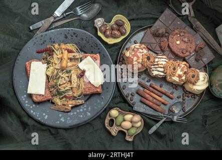 Spaghetti à la saucisse au fromage sur le pain dans une assiette en céramique noire servie avec des rouleaux de cannelle, des barres de chocolat noir, des biscuits et des morceaux en pla en céramique noire Banque D'Images