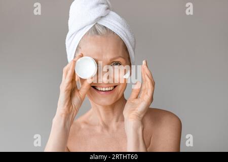 Belle femme mature tenant un pot avec une crème hydratante à la main, couvrant un œil avec un produit de soin de la peau Banque D'Images