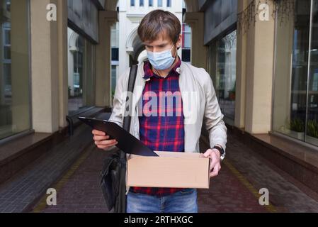 Portrait d'un jeune homme dans un masque médical, protecteur, avec une boîte de choses, qui a perdu son emploi. Banque D'Images