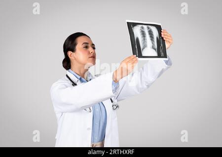Jeune médecin latine tenant et regardant la radiographie thoracique, debout sur fond gris de studio Banque D'Images