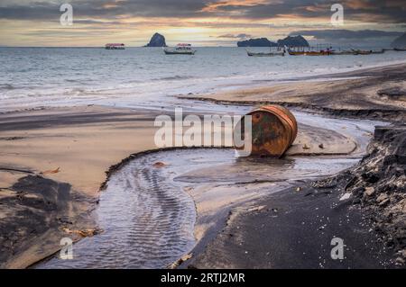 Le baril de pétrole rouillé sur une plage en partie noire illustre la pollution de l'environnement par les déversements de pétrole. Bateaux Longtail en arrière-plan au coucher du soleil Banque D'Images