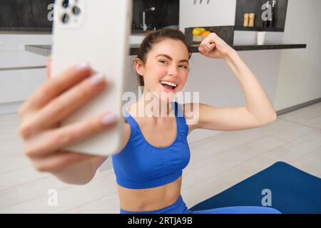 Portrait de fille de fitness active, prend selfie sur smartphone pendant l'entraînement à la maison, posant sur le tapis de fitness, montre ses muscles forts et rit Banque D'Images