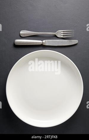 Assiette en céramique blanche et fourchette à couverts et couteau sur fond de béton noir. Gabarit pour afficher les aliments sur le menu Banque D'Images