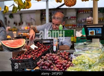 Homme servant dans le stand du marché avec raisins frais, cerises et pastèque sur l'affichage par la machine de poids Banque D'Images