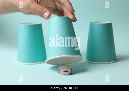 Femme montrant des pièces de monnaie sous la tasse sur fond bleu clair, closeup. Jeu Thimblerig Banque D'Images