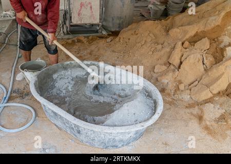 Travailleur mélanger manuellement la poudre de ciment sur le chantier de construction, en utilisant une houe pour mélanger la poudre de ciment, le sable, les pierres dans le bassin pour mélanger le ciment. Banque D'Images