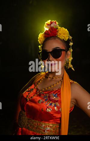 Une danseuse sundanaise pose devant une lumière brillante tout en portant un costume orange avec des lunettes de soleil et des fleurs sur la tête la nuit Banque D'Images