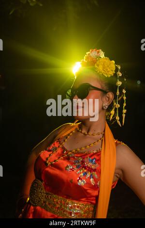 Une danseuse sundanaise pose devant une lumière brillante tout en portant un costume orange avec des lunettes de soleil et des fleurs sur la tête la nuit Banque D'Images