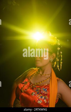 Une danseuse sundanaise pose devant une lumière brillante tout en portant un costume orange avec des lunettes de soleil et des fleurs sur la tête la nuit Banque D'Images