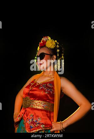 Une danseuse sundanaise pose devant une lumière brillante tout en portant un costume orange avec des lunettes de soleil et des fleurs sur la tête la nuit Banque D'Images