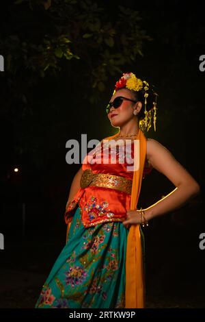 Une danseuse sundanaise pose devant une lumière brillante tout en portant un costume orange avec des lunettes de soleil et des fleurs sur la tête la nuit Banque D'Images