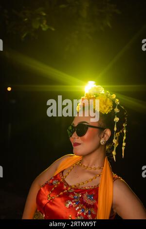 Une danseuse sundanaise pose devant une lumière brillante tout en portant un costume orange avec des lunettes de soleil et des fleurs sur la tête la nuit Banque D'Images