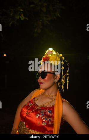 Une danseuse sundanaise pose devant une lumière brillante tout en portant un costume orange avec des lunettes de soleil et des fleurs sur la tête la nuit Banque D'Images