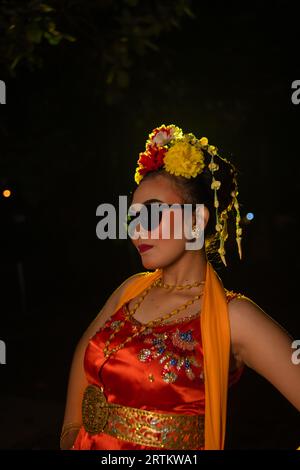 Une danseuse sundanaise pose devant une lumière brillante tout en portant un costume orange avec des lunettes de soleil et des fleurs sur la tête la nuit Banque D'Images