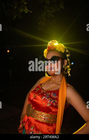 Une danseuse sundanaise pose devant une lumière brillante tout en portant un costume orange avec des lunettes de soleil et des fleurs sur la tête la nuit Banque D'Images