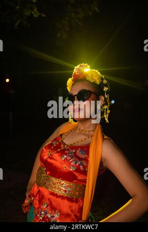 Une danseuse sundanaise pose devant une lumière brillante tout en portant un costume orange avec des lunettes de soleil et des fleurs sur la tête la nuit Banque D'Images