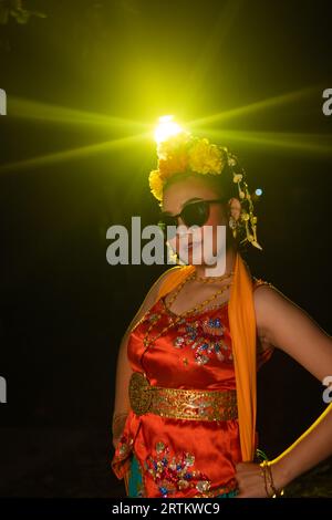 Une danseuse sundanaise pose devant une lumière brillante tout en portant un costume orange avec des lunettes de soleil et des fleurs sur la tête la nuit Banque D'Images