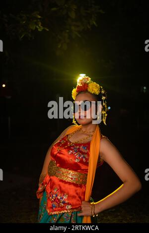 Une danseuse sundanaise pose devant une lumière brillante tout en portant un costume orange avec des lunettes de soleil et des fleurs sur la tête la nuit Banque D'Images