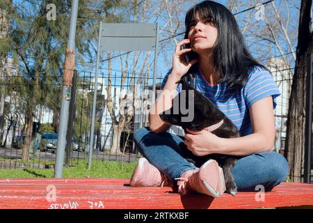 Jeune femme vénézuélienne latine aux cheveux noirs, vêtue de bleu, assise dans le parc avec son chien attentif parlant au téléphone au coucher du soleil, concept de technologie, Banque D'Images