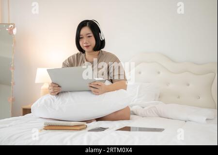 Une jolie femme asiatique de grande taille appréciant son temps libre à la maison, portant des écouteurs, écoutant de la musique ou regardant un film sur son ordinateur portable au lit. Banque D'Images