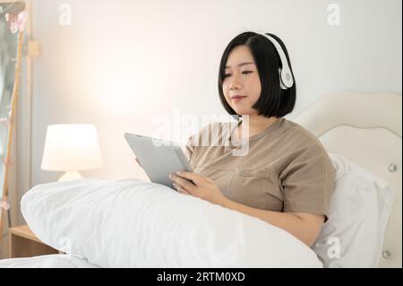 Une belle femme asiatique de grande taille dans des vêtements décontractés portant des écouteurs et utilisant sa tablette numérique tout en se relaxant sur son lit dans sa chambre. Banque D'Images