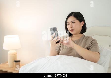 Une belle femme asiatique de grande taille dans des vêtements confortables utilisant son smartphone, bavardant avec ses amis tout en se relaxant sur son lit dans sa chambre. Personnes an Banque D'Images
