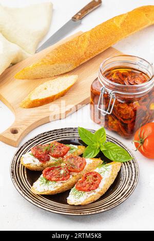 Pain tranché, un bocal ouvert et des sandwichs prêts à l'emploi avec des tomates séchées, du fromage et de l'aneth dans une assiette sur la table. Banque D'Images