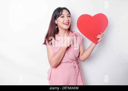 Femme asiatique excitée vêtue de rose, pointant vers l'espace de copie à côté d'elle tout en tenant un grand papier rouge en forme de coeur, isolé par fond blanc Banque D'Images