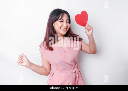 Une jeune femme asiatique avec une expression heureuse réussie portant un chemisier rose et tenant un papier rouge en forme de coeur, isolé par fond blanc Banque D'Images