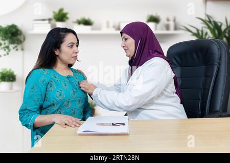 Médecin musulman féminin portant le hijab tenant stéthoscope vérifier le battement de coeur de jeune femme indienne dans la clinique moderne. Banque D'Images