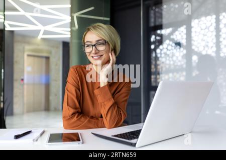 Portrait de la jeune femme d'affaires réussie à l'intérieur du bureau au travail, la travailleuse est rêveuse et pensive souriante et regardant par la fenêtre, la femme pense tout en travaillant avec un ordinateur portable. Banque D'Images