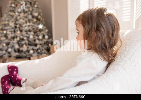 Une petite fille contrariée ne veut pas être photographiée, se détourne de l'appareil photo, couvre l'objectif de sa main. Nocif, vilain enfant assis en blanc Banque D'Images