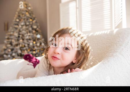 Drôle petite fille se trouve dans une chaise blanche douce, trompant autour, faisant des visages près de l'arbre de Noël. Enfant dans une atmosphère chaleureuse à la maison. interi festif Banque D'Images