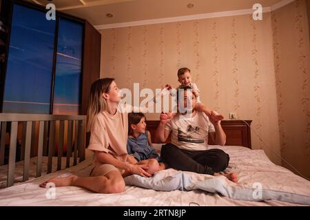 Portrait drôle de famille joyeuse diverse sur le lit le matin. Mère élégante embrasse sa fille, père avec des lunettes tient le petit fils sur les épaules. Parents Banque D'Images