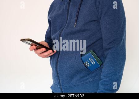 Homme en sweat-shirt bleu avec smartphone à la main et passeport étranger ukrainien dans la poche sur fond blanc Banque D'Images