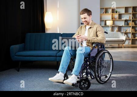 Bel homme handicapé assis en fauteuil roulant avec un ordinateur portable sur les genoux et regardant la caméra Banque D'Images