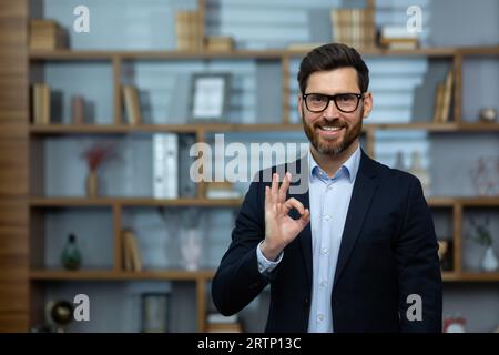 OK signe, homme d'affaires montre le signe d'approbation à la caméra, homme souriant dans le costume d'affaires à l'intérieur du bureau au lieu de travail, investisseur financier expérimenté dans le bureau. Banque D'Images