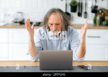 Déçu caucasien gris cheveux mature homme, retraité, mentor financier, s'asseoir à une table dans la cuisine, regarde confusionnement à un ordinateur portable, est perplexe par les nouvelles ou le résultat du travail, gesturant les mains Banque D'Images